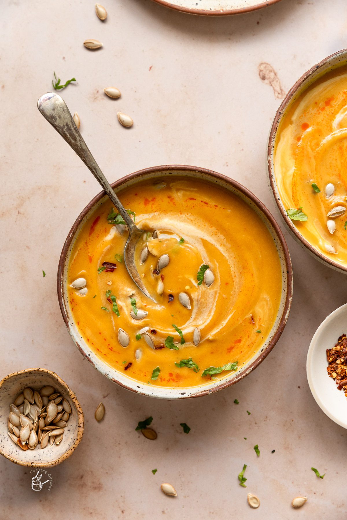 Three curried pumpkin soup bowls on a dining table next to small bowl of pumpkin seeds and chilli flakes.