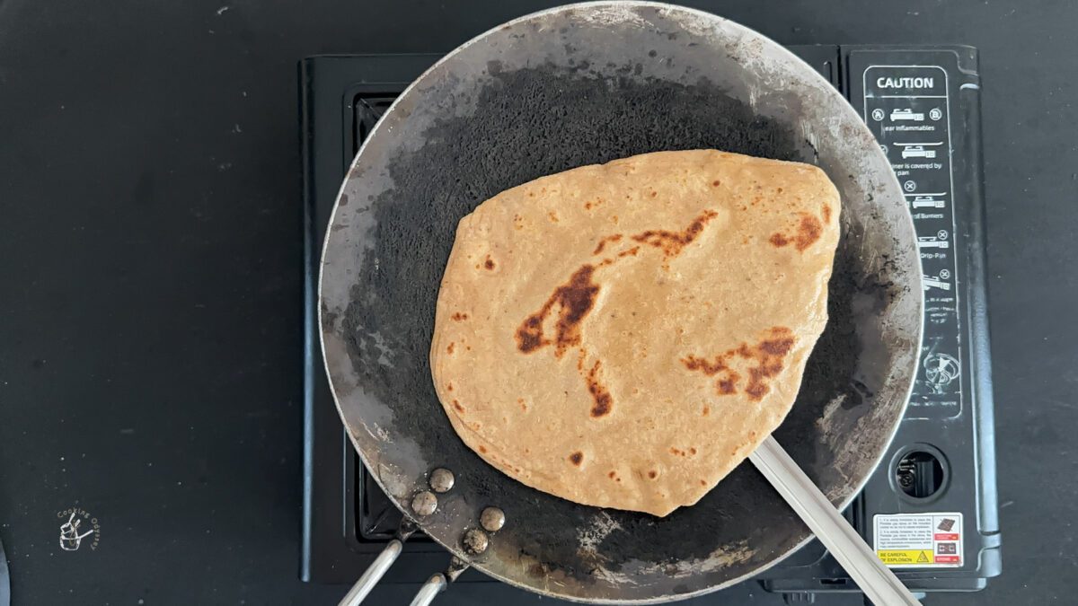 A female hand is picking up a cooked ajwain paratha from the tawa using a spatula.