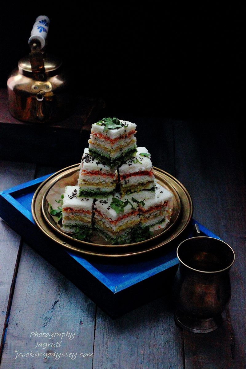 a stack of three layered dhokla. first layer has a red chutney, nect a yellow chutney and finally green chutney