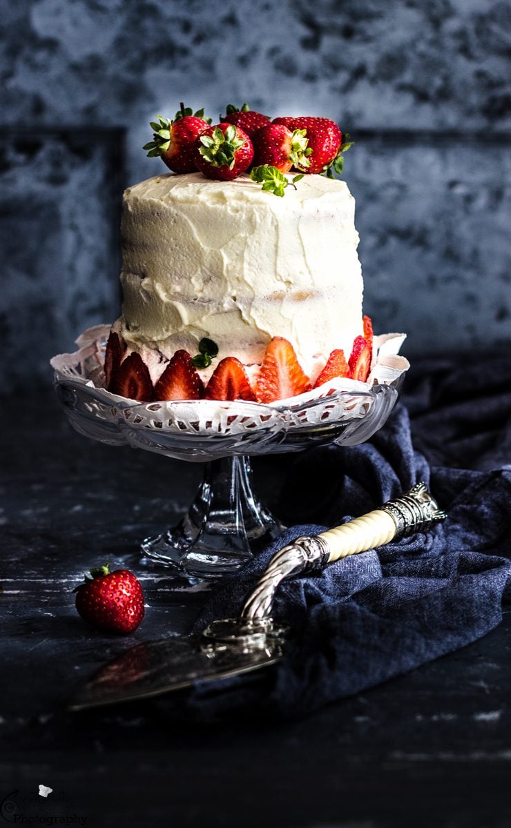 3 layered sponge cake covered in whipped cream and strawberry slices on a cake stand against a blue background.