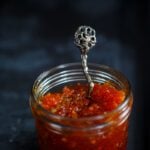 a glass jar filled with red pepper chutney with a decorative spoon to serve