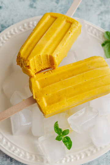 Two mango popsicles on a round plate with a bite taken out from one.
