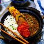 a black bowl filled with rice on one side and katsu curry sauce on the other and topped with vegetable katsu pieces and chopsticks