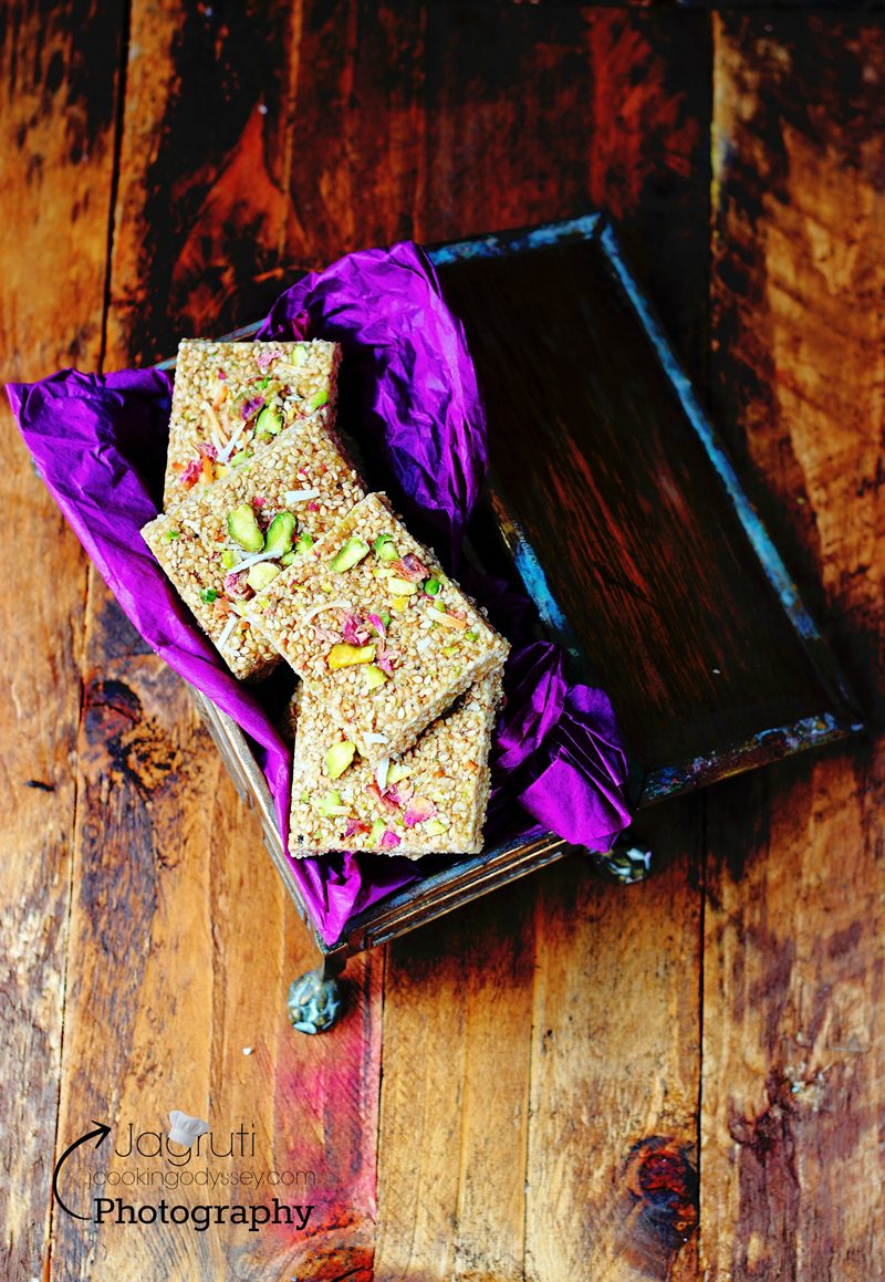 a small tray layered with a purple cloth. on top lays sesame seed brittle pieces which are topped with nuts and rose petals