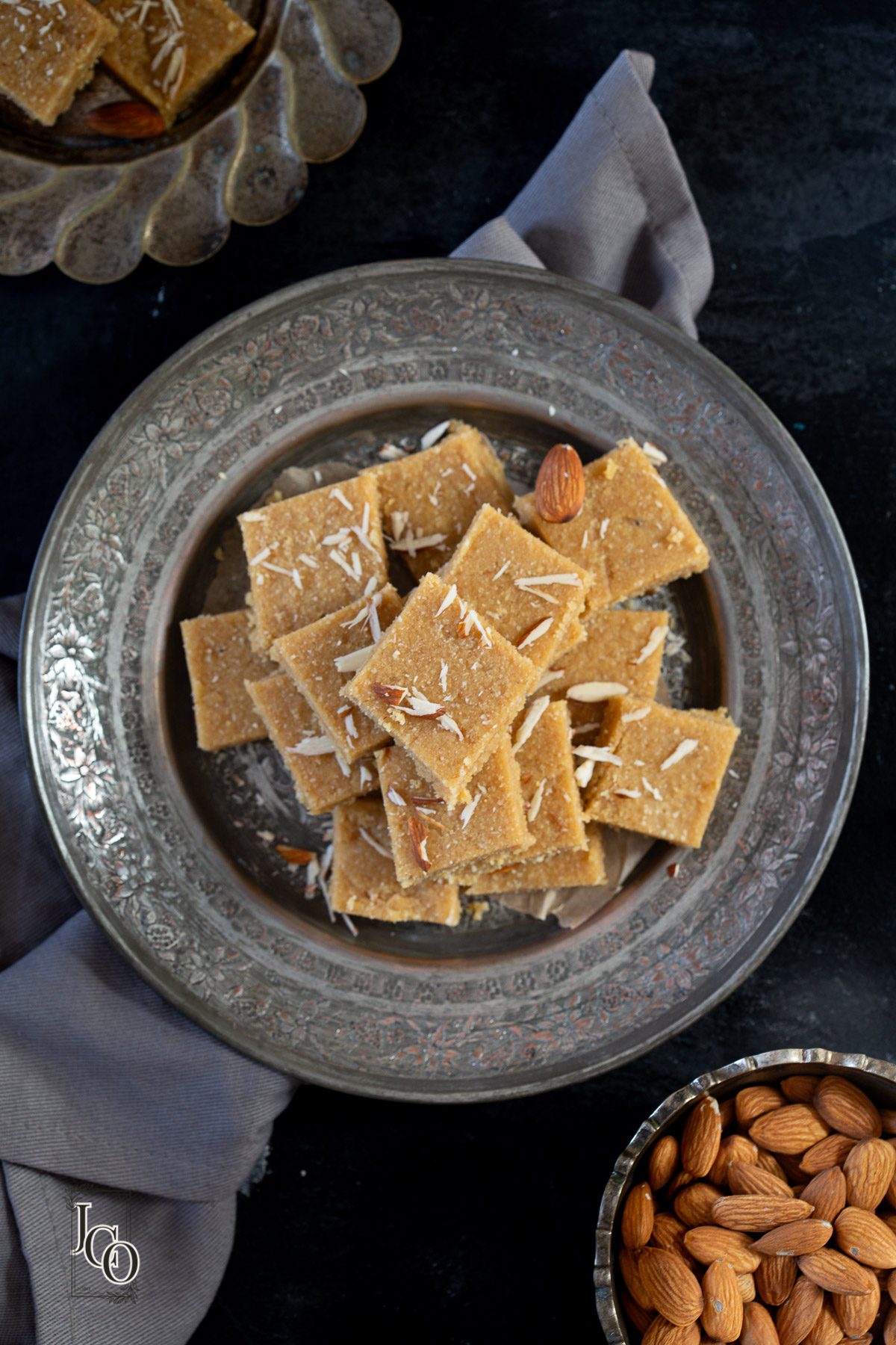 Almond sukhdi served in a metal serving plate, next to a bowl filled with raw almonds.