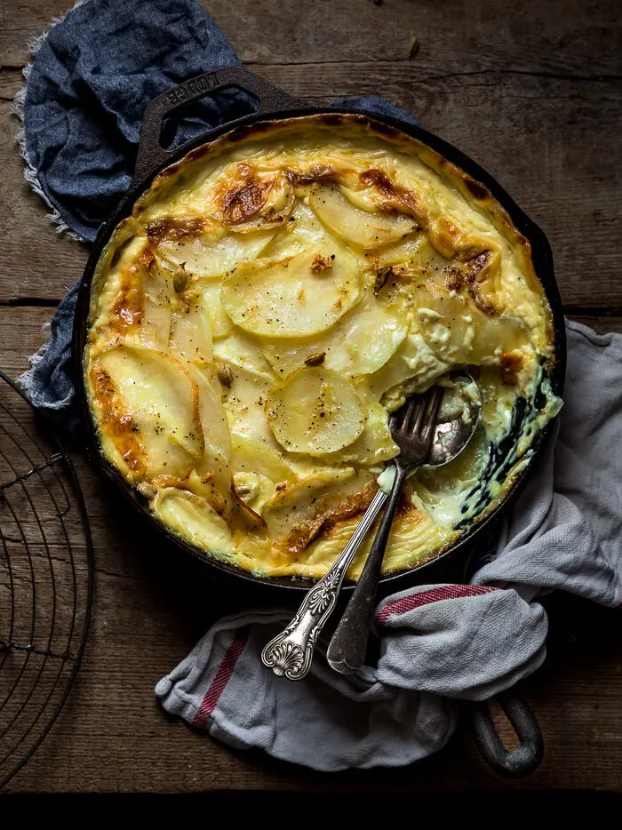 cardamom dauphinoise potatoes are in a cast iron skillet next to serving spoon