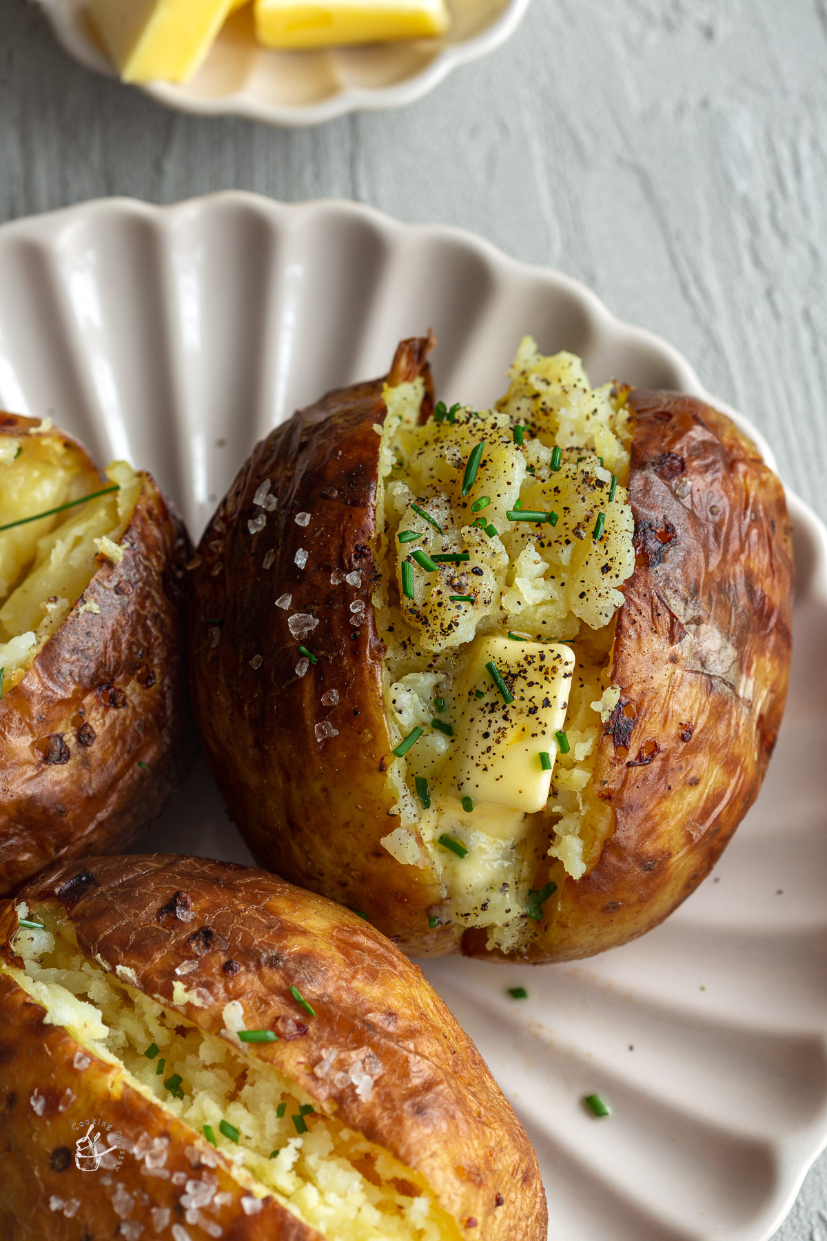 three baked potatoes on a beige fluted plate. the potatoes are filled with butter and chives