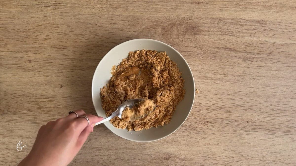 a female hand is mixing butter and crushed biscuits in a bowl with a spoon