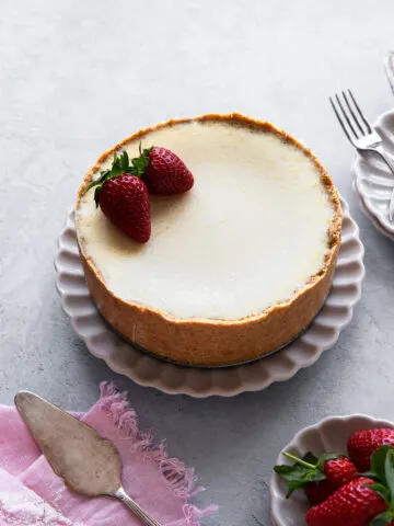 whole eggless baked cheesecake served on a white flute plate next to strawberries bowl and pink kitchen napkin