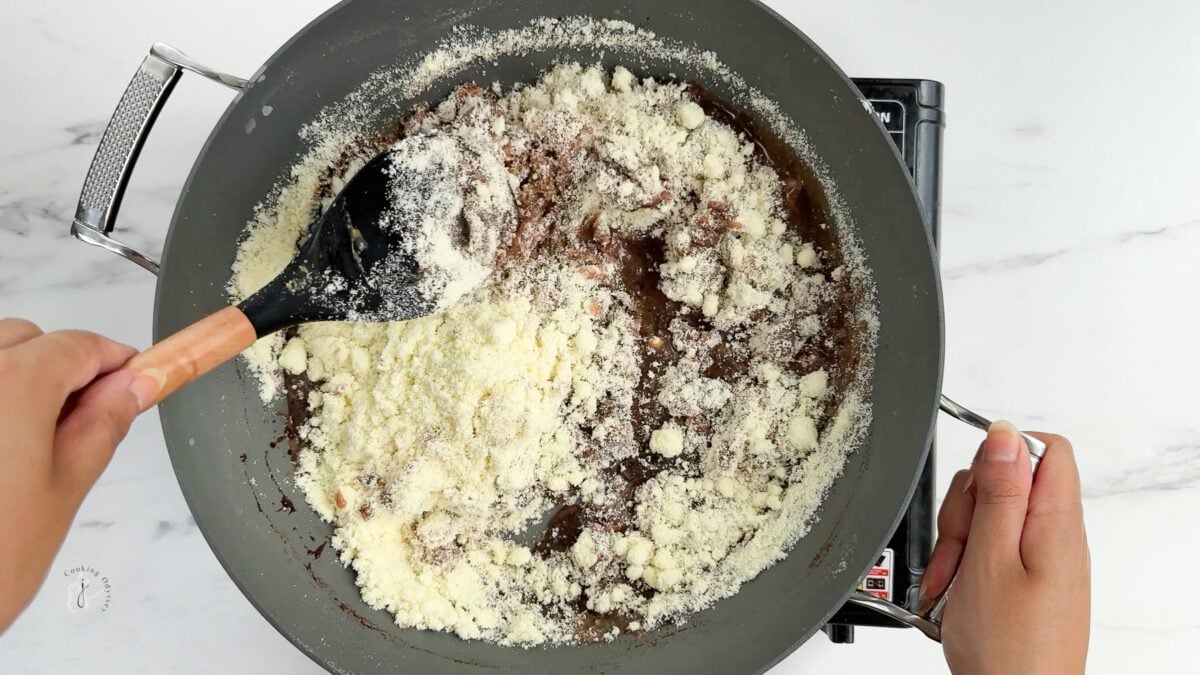 a female hands are mixing milk powder and chocolate condenced milk in the pan to make chocolate barfi 