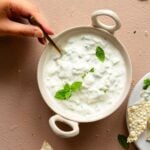 A female hand is serving cucumber raita using a spoon.