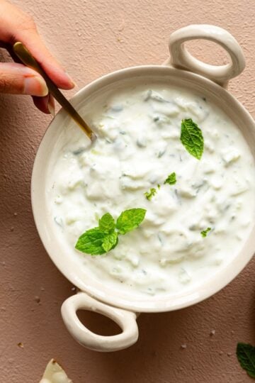 A female hand is serving cucumber raita using a spoon.