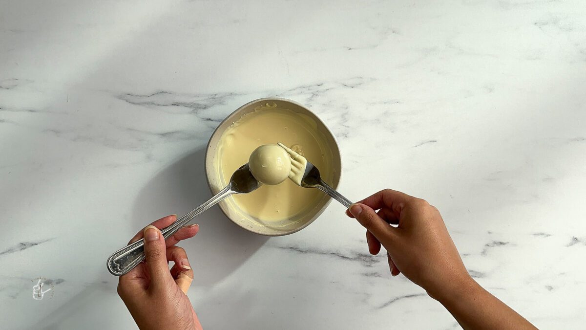 Female hands are coating a gulab jamun in melted white chocolate using two forks