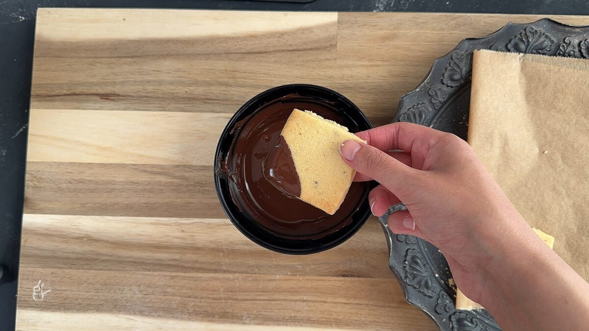 a female hand is dipping a barfi biscuit in dark melted chocolate