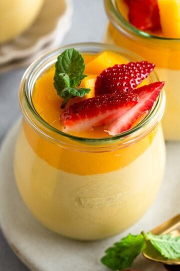 Two jars of eggless mango mousse on a round marble chopping board next to dessert spoons.