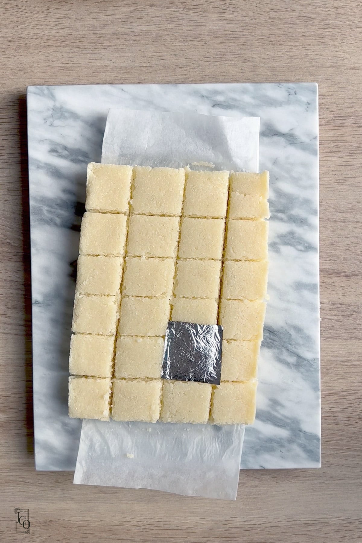 Twenty four square pieces of coconut barfi laid on a marble slab. 
