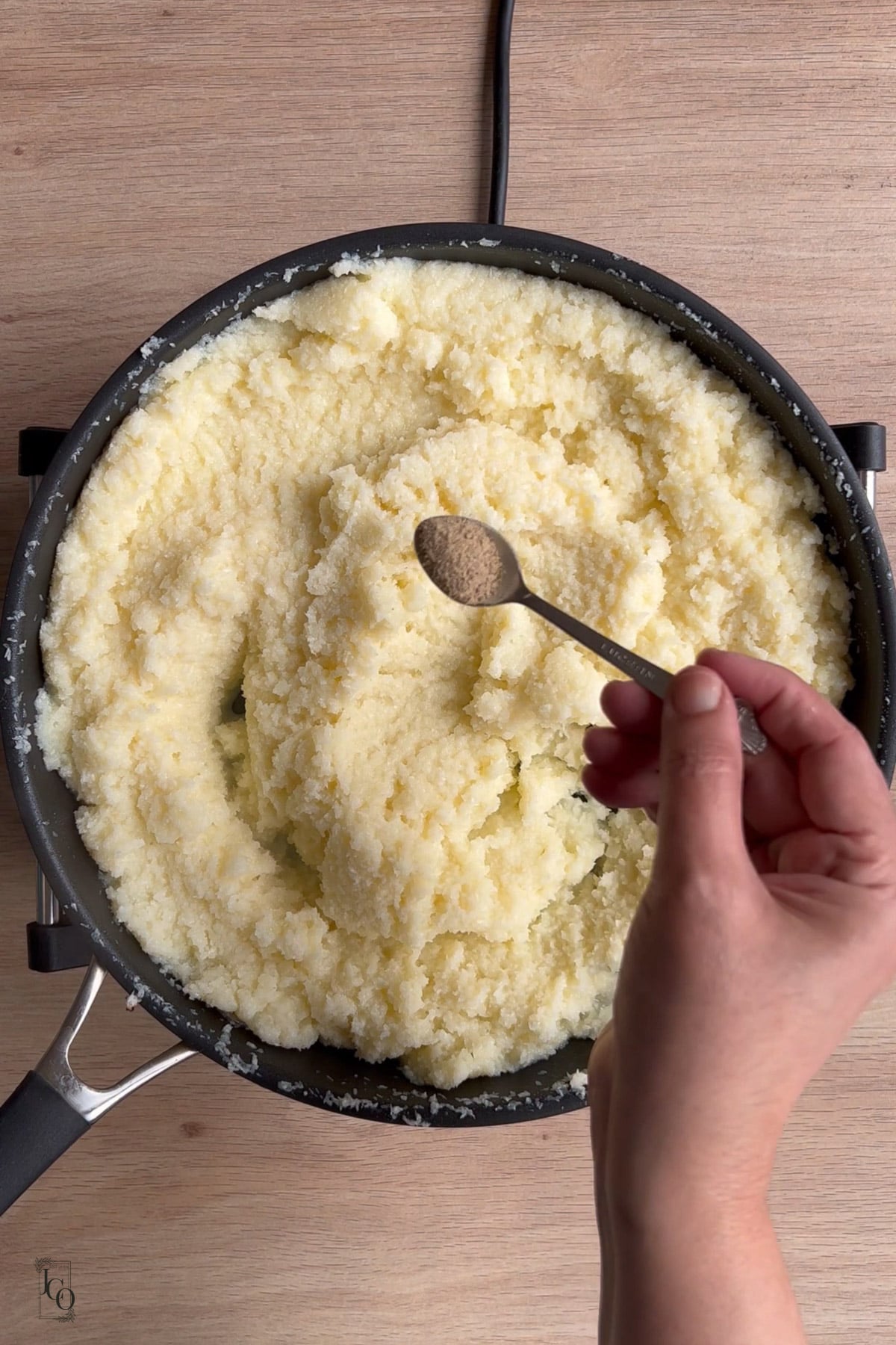 A female hand is holding a spoon of cardamom powder above the cooked coconut mixture in a pan. 
