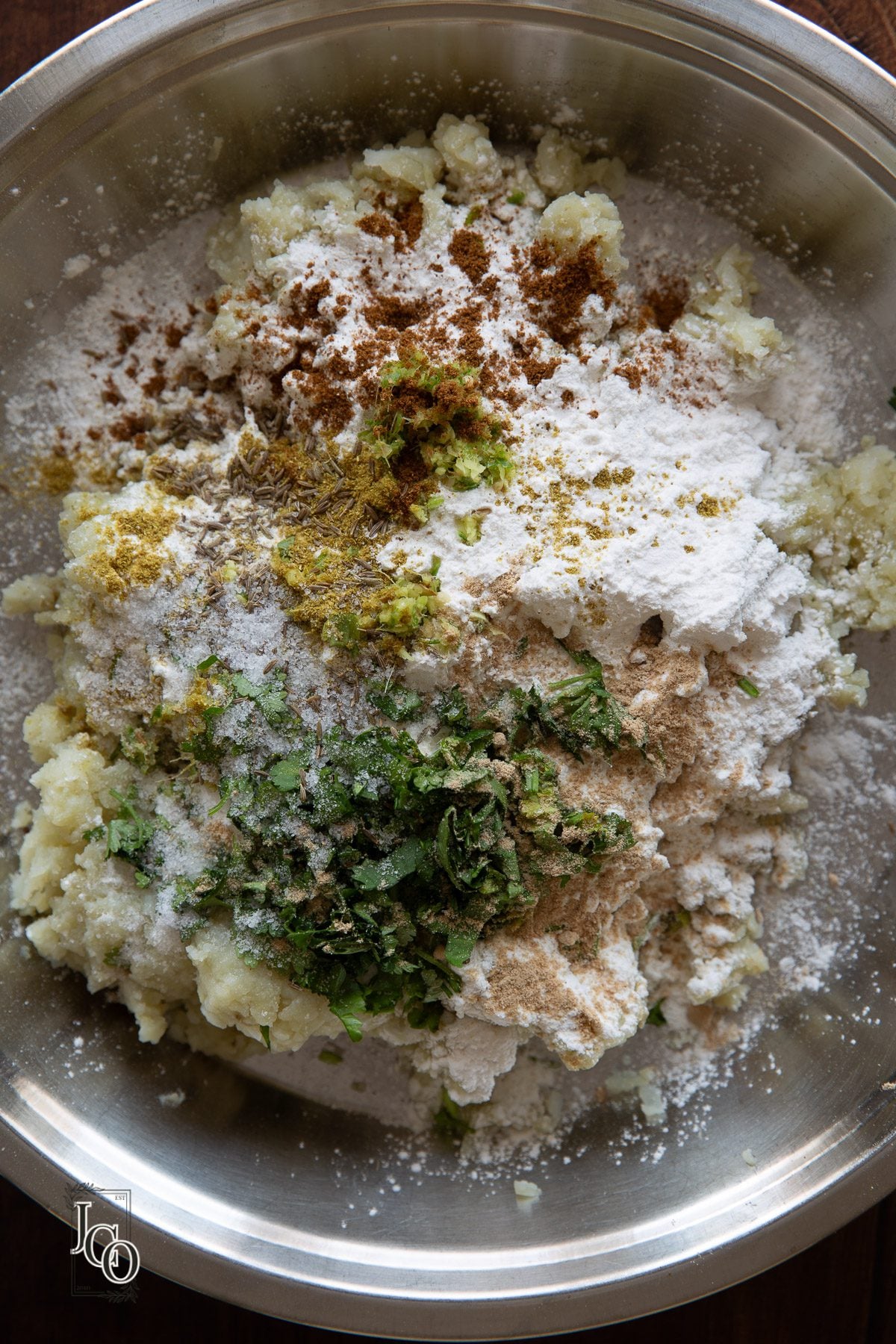 Spices, herbs and corn flour added to the grated potatoes in a pan.