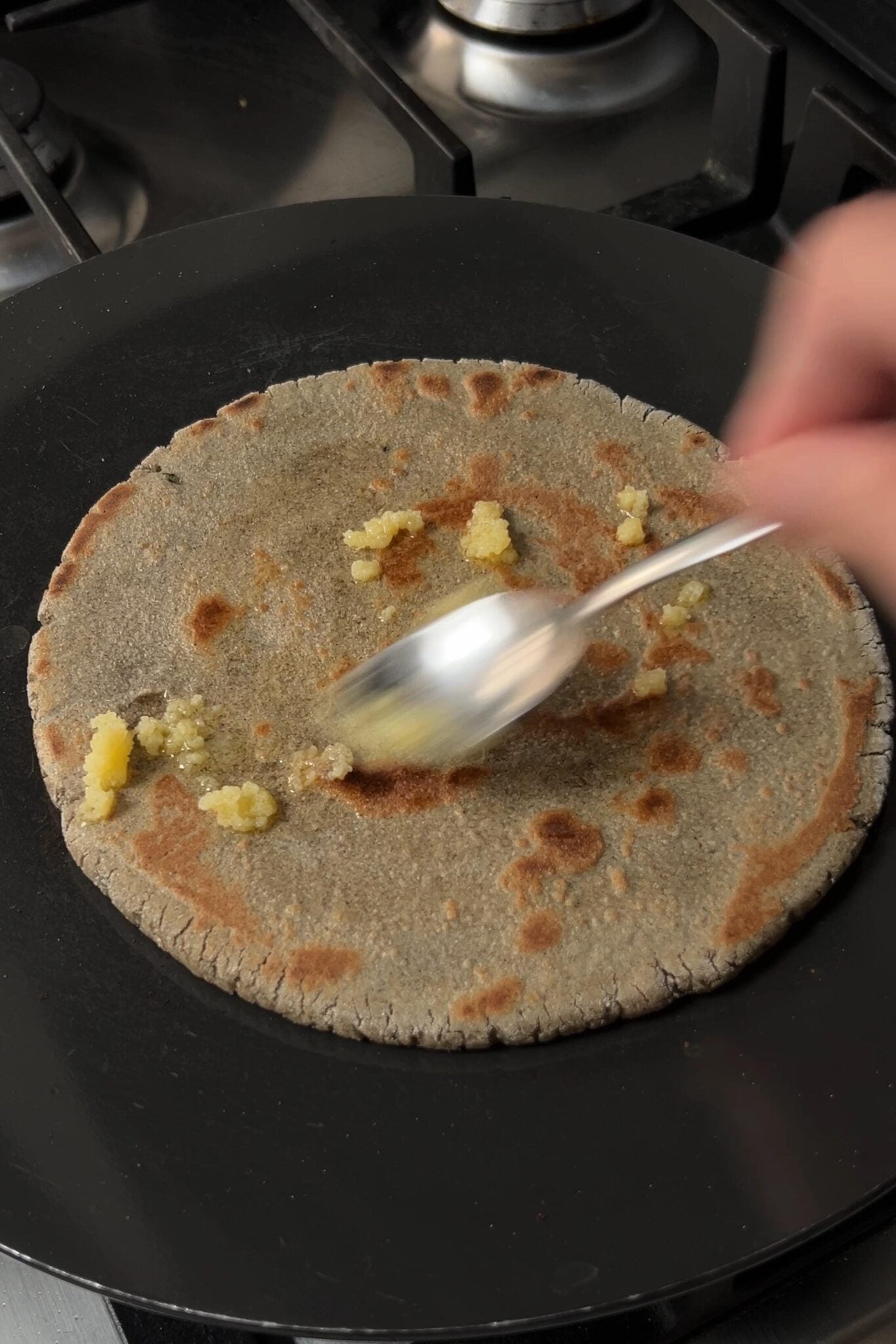 A female hand is applying ghee on a flatbread using a spoon on hot griddle.