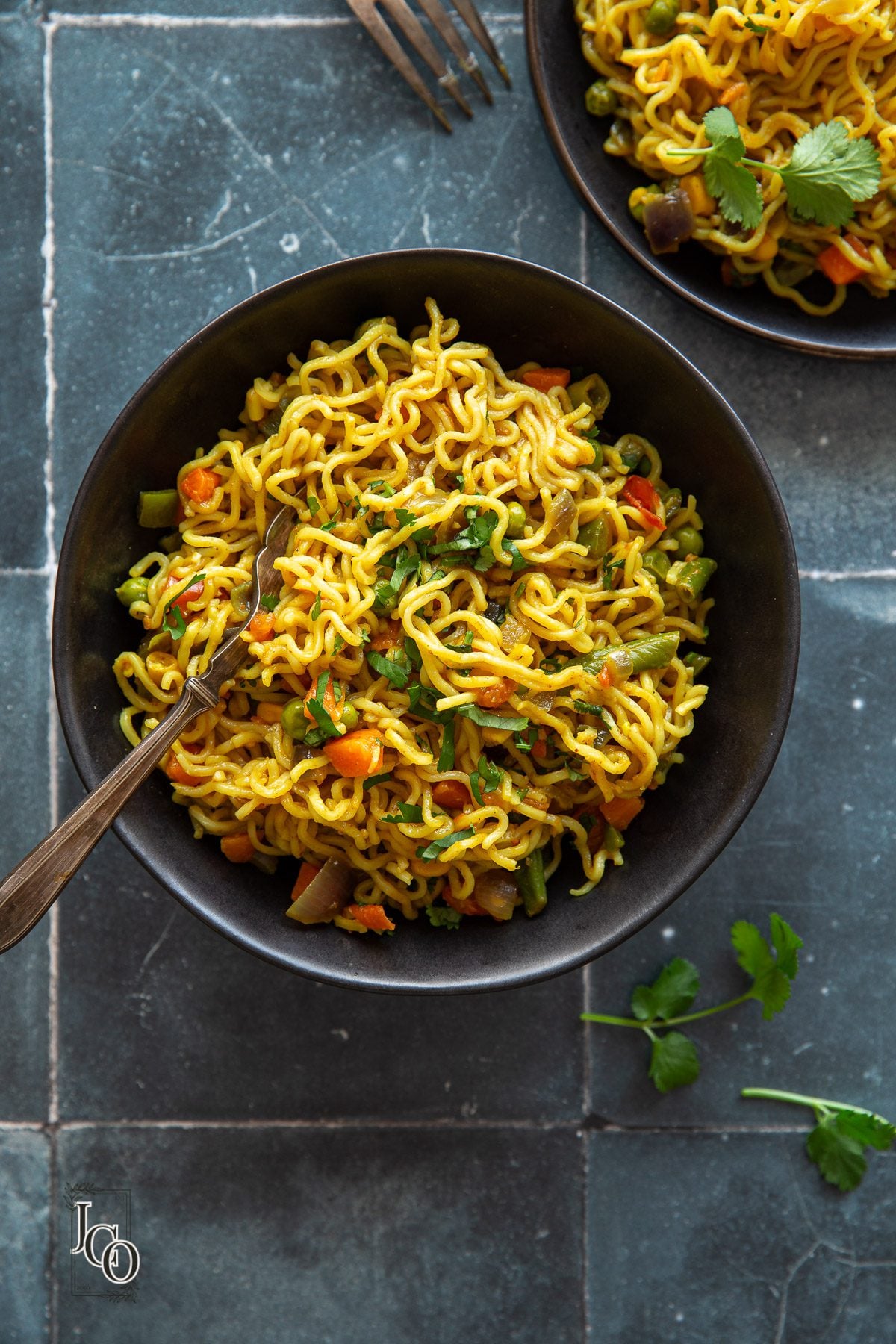 Masala maggi noodles served in two black ceramic bowls, garnished with chopped coriander on top.