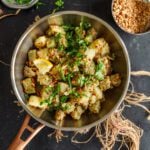 Sweet potato nu shaak in a pan, next to a bowl of chopped herbs and crushed peanuts.