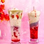 Two tall glasses of pistachio rose falooda placed near the flaower vase on the table.