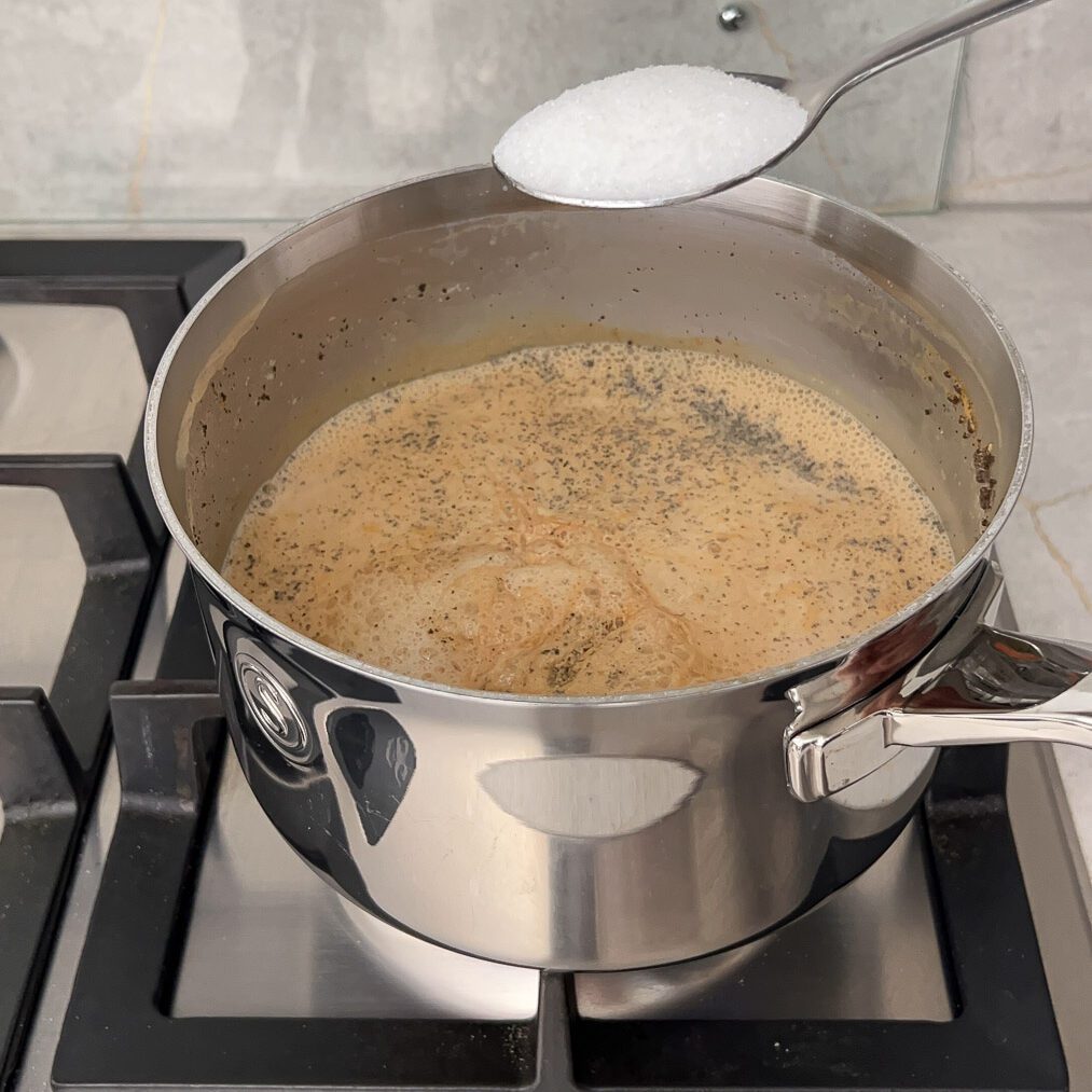 Sugar being added using a spoon in boiling chai in the saucepan over the stovetop.