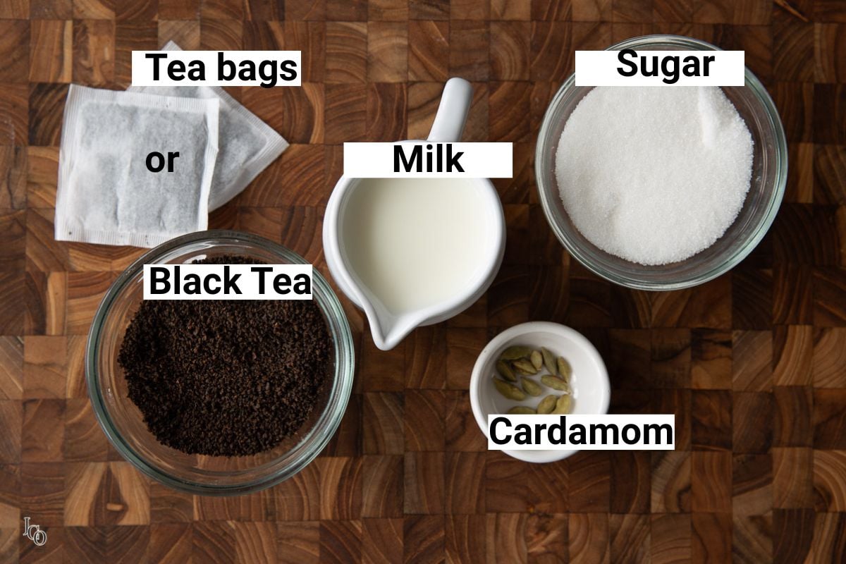 Prepared and labeled ingredients of doodh patti chai on brown surface like milk, tea leaves, sugar and cardamom pods. 