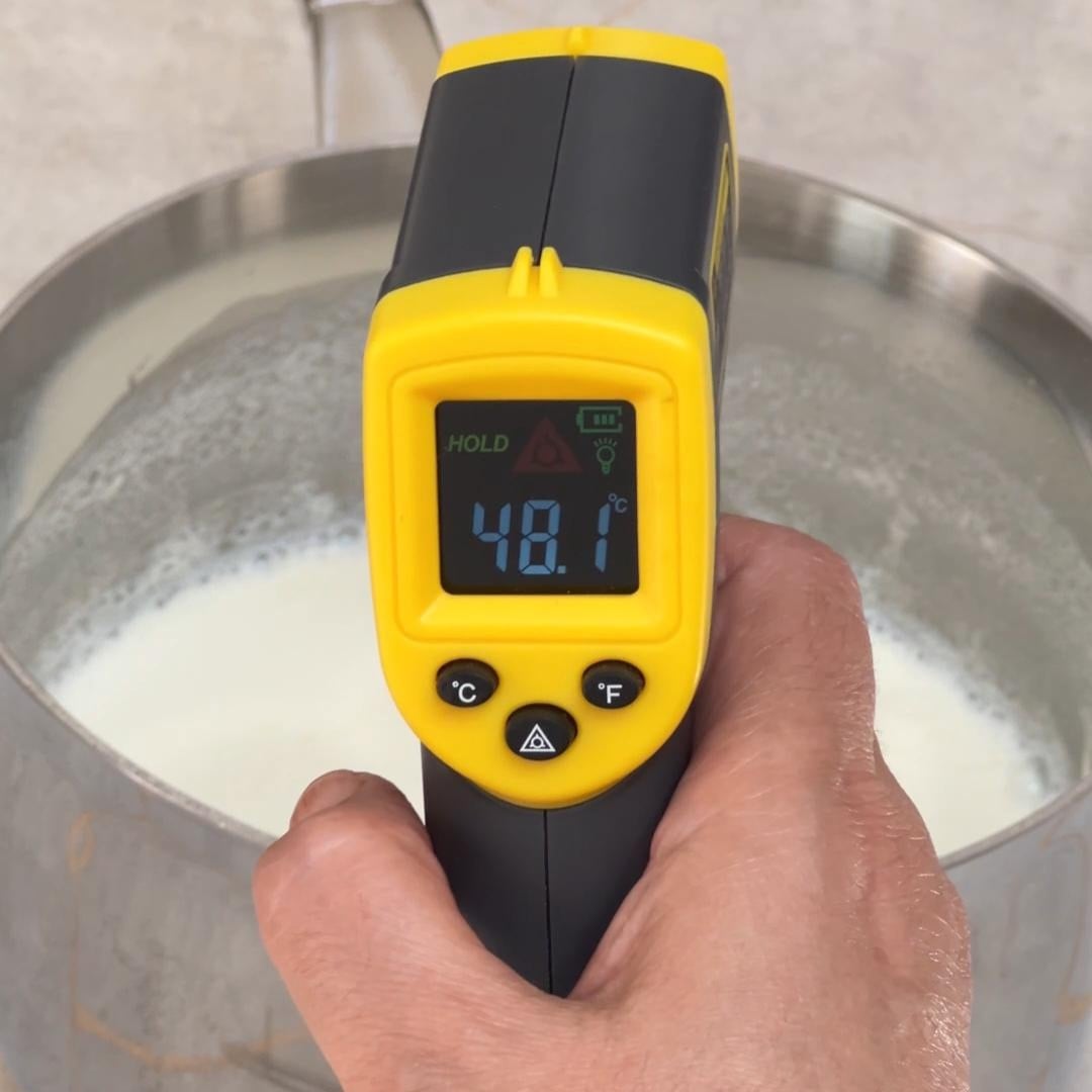 A female hand is pointing a thermometer which shows 48C at milk in a saucepan. 