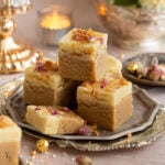Two layered barfi squares placed on a serving tray, background candles lit in a glass candle holder.