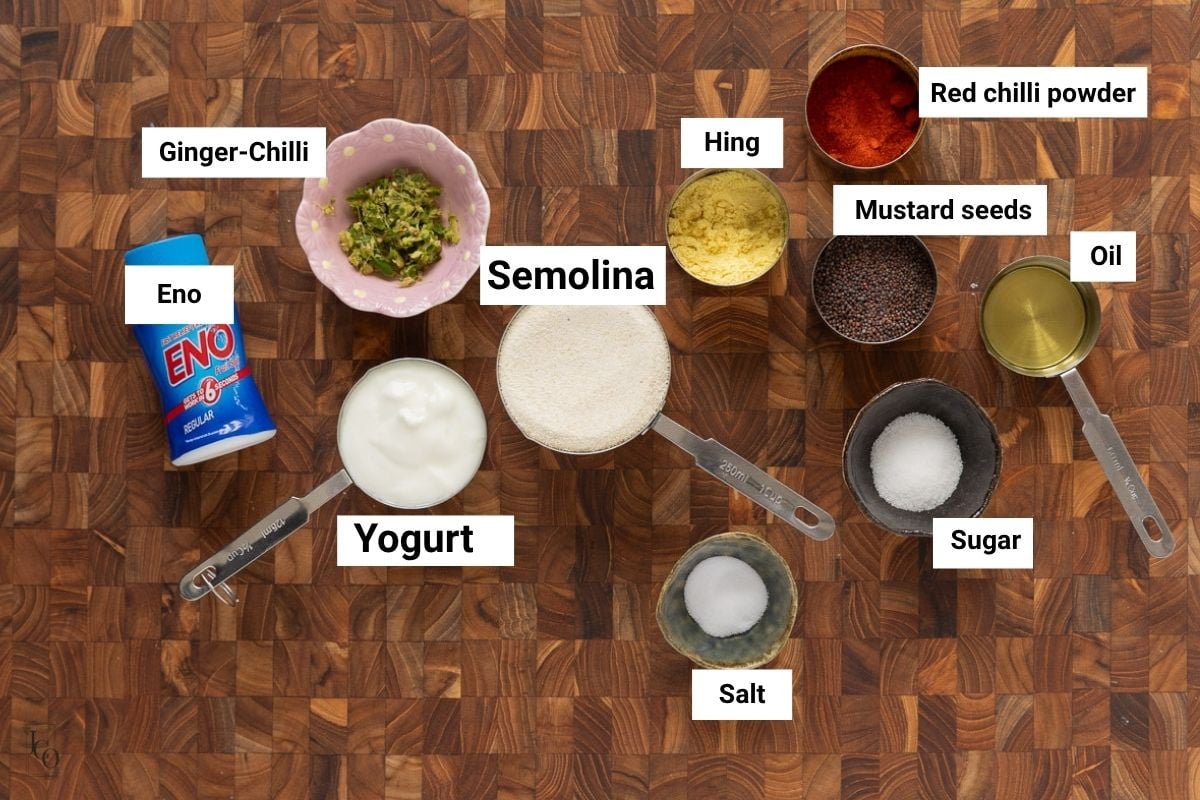 Semolina, yogurt, eno, spices, sugar and oil in small bowls on brown backdrop.