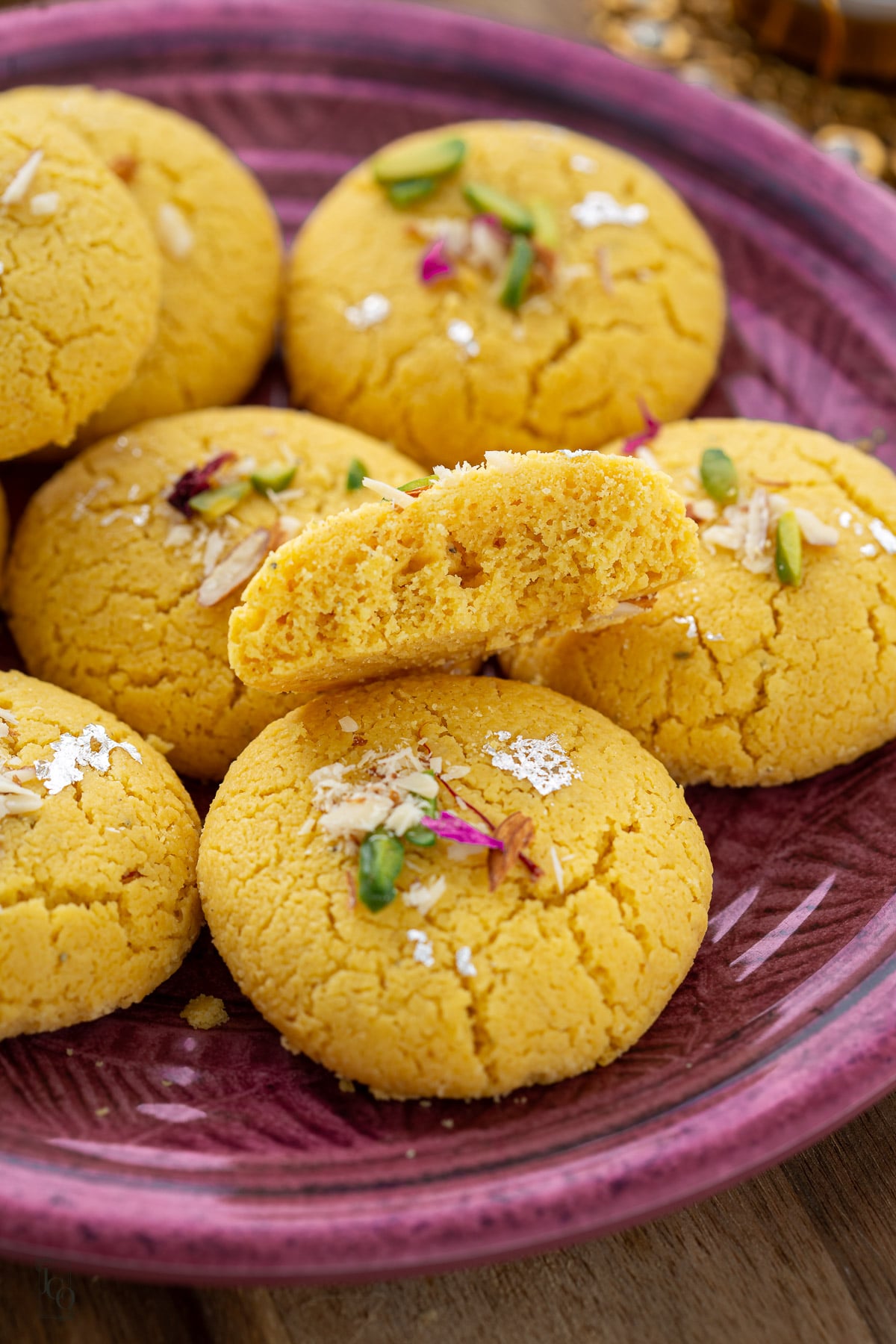 A purple plate filled with round, yellow besan nankhatai topped with nuts, petals, and silver foil, with one cookie broken in half to reveal a perfectly baked texture.