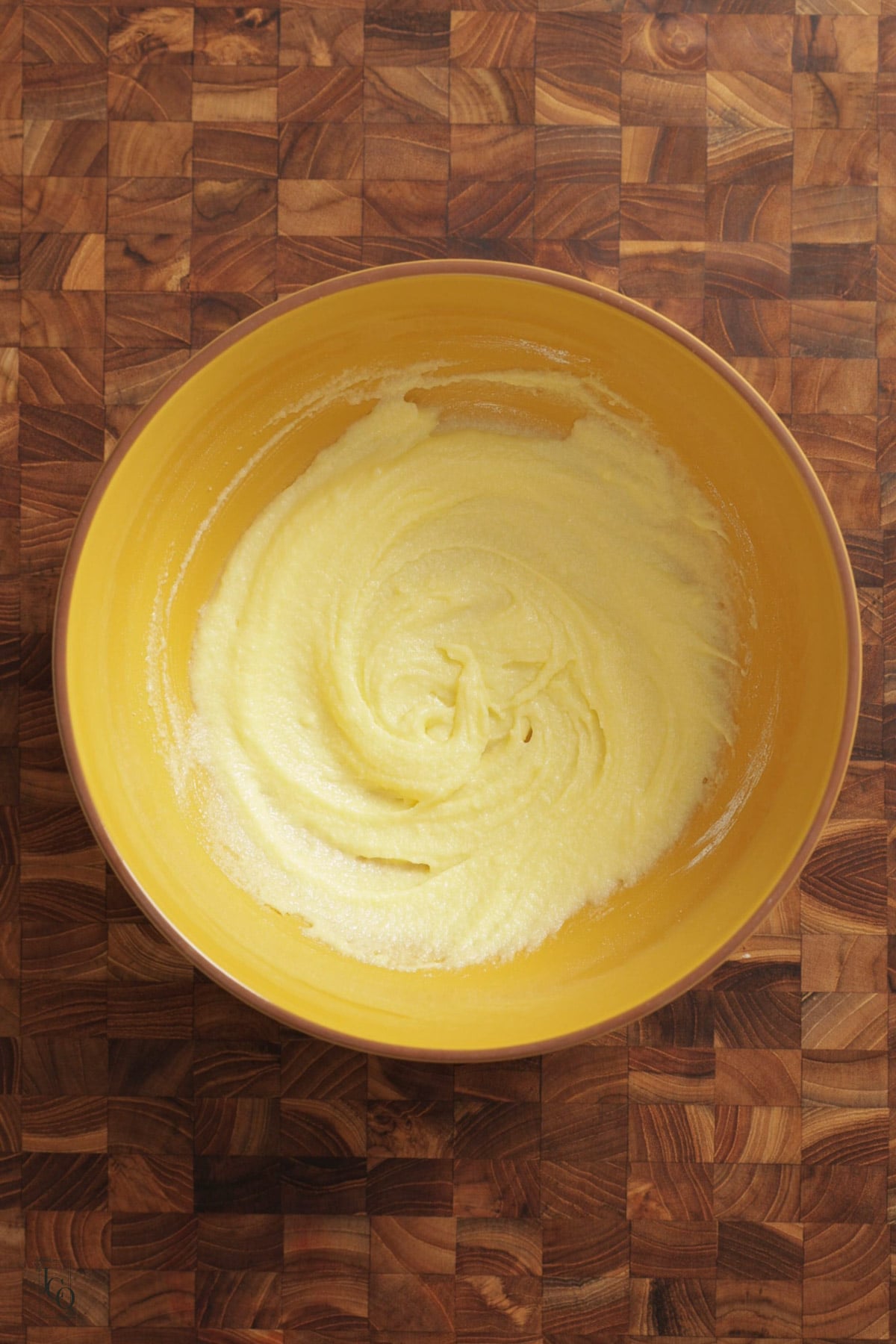 A yellow mixing bowl containing creamy, pale yellow mixture of ghee and caster sugar on a wooden surface.