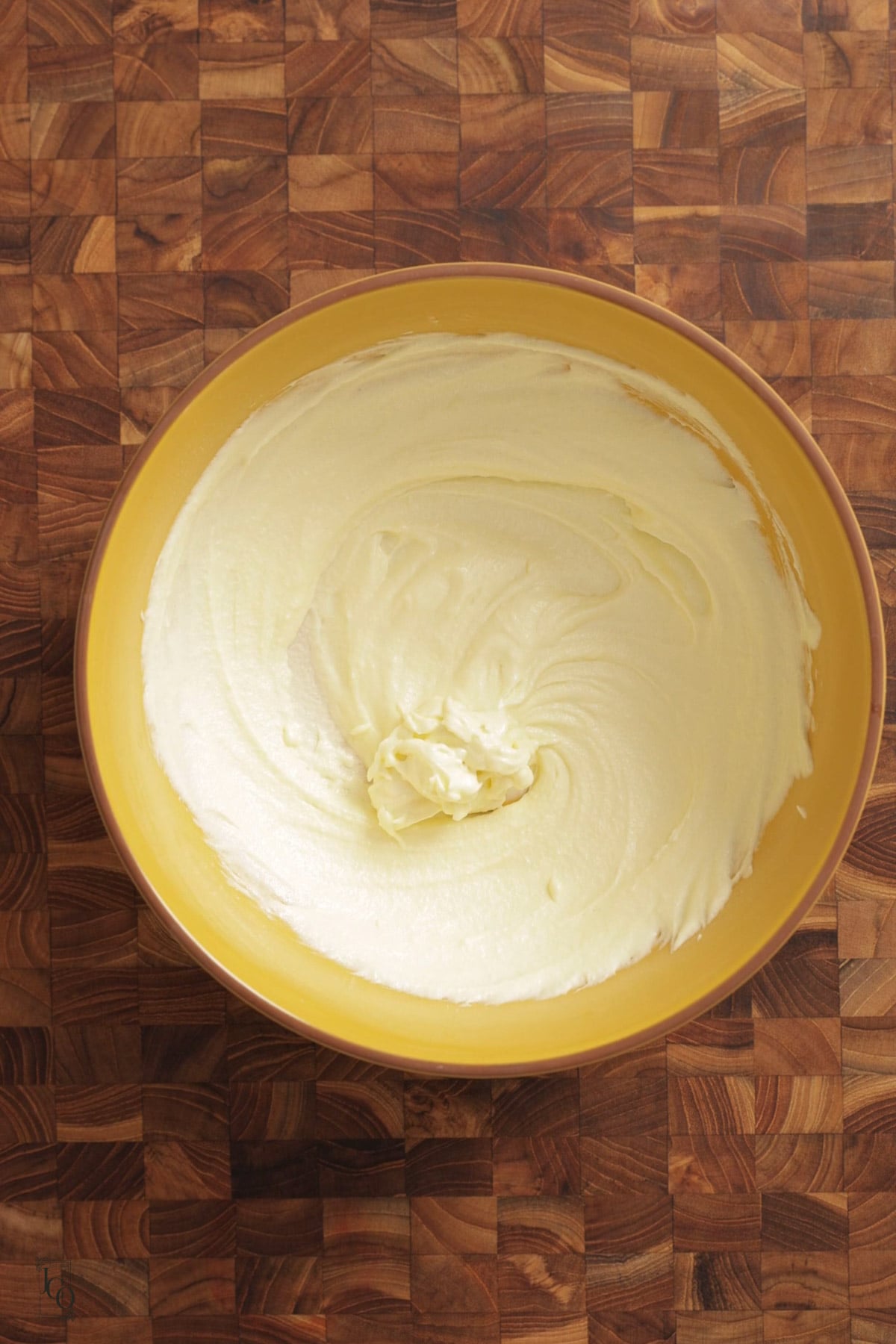 Airy and creamy white ghee and sugar mixture in a yellow mixing bowl on a wooden cutting board.