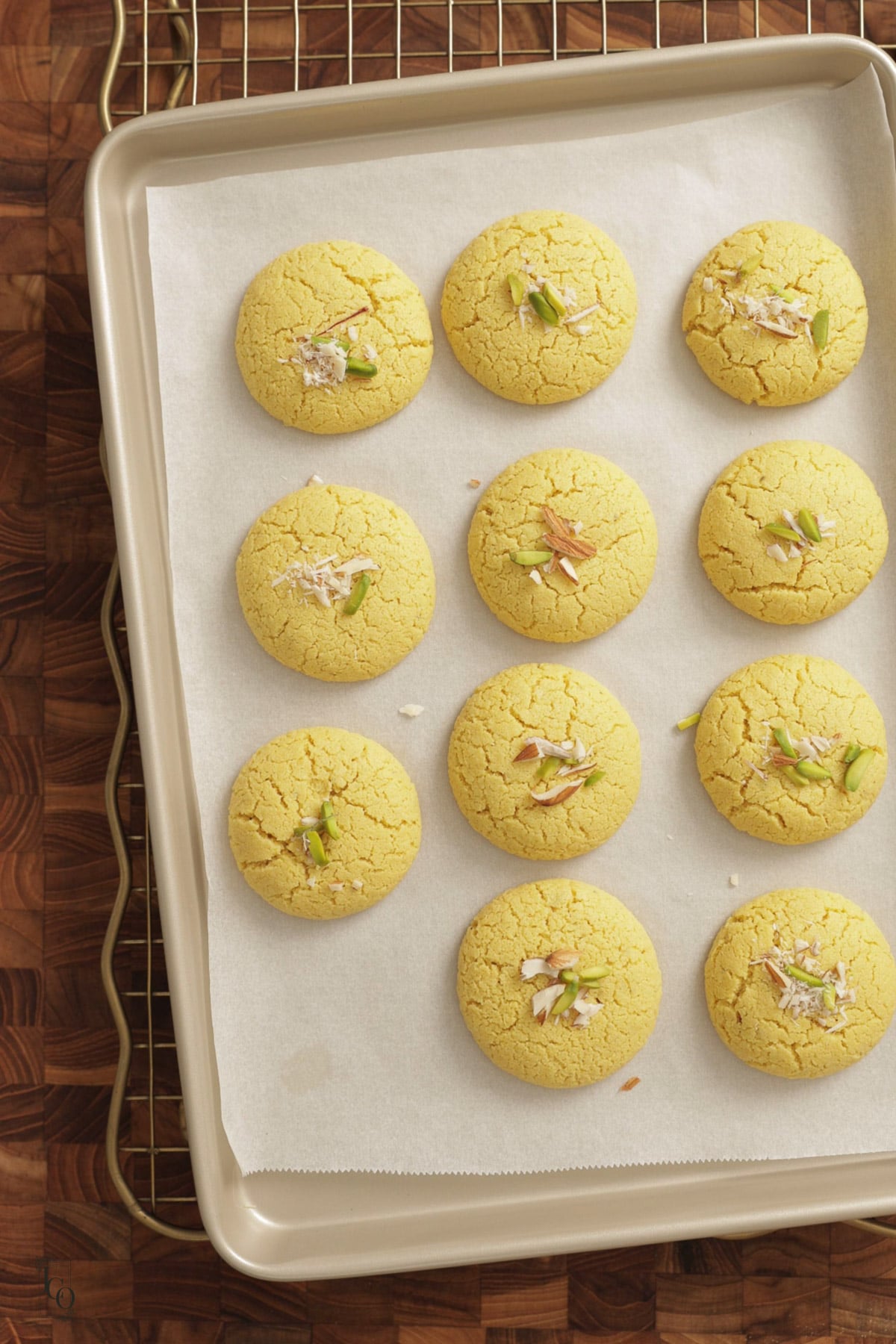 A baking tray lined with greaseproof paper holds eleven round, yellow besan nankhatai biscuits topped with slivered nuts.