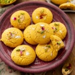 A purple plate piled with round, golden-yellow besan nankhatai biscuits topped with slivered nuts, dried rose petals, and edible silver leaf.