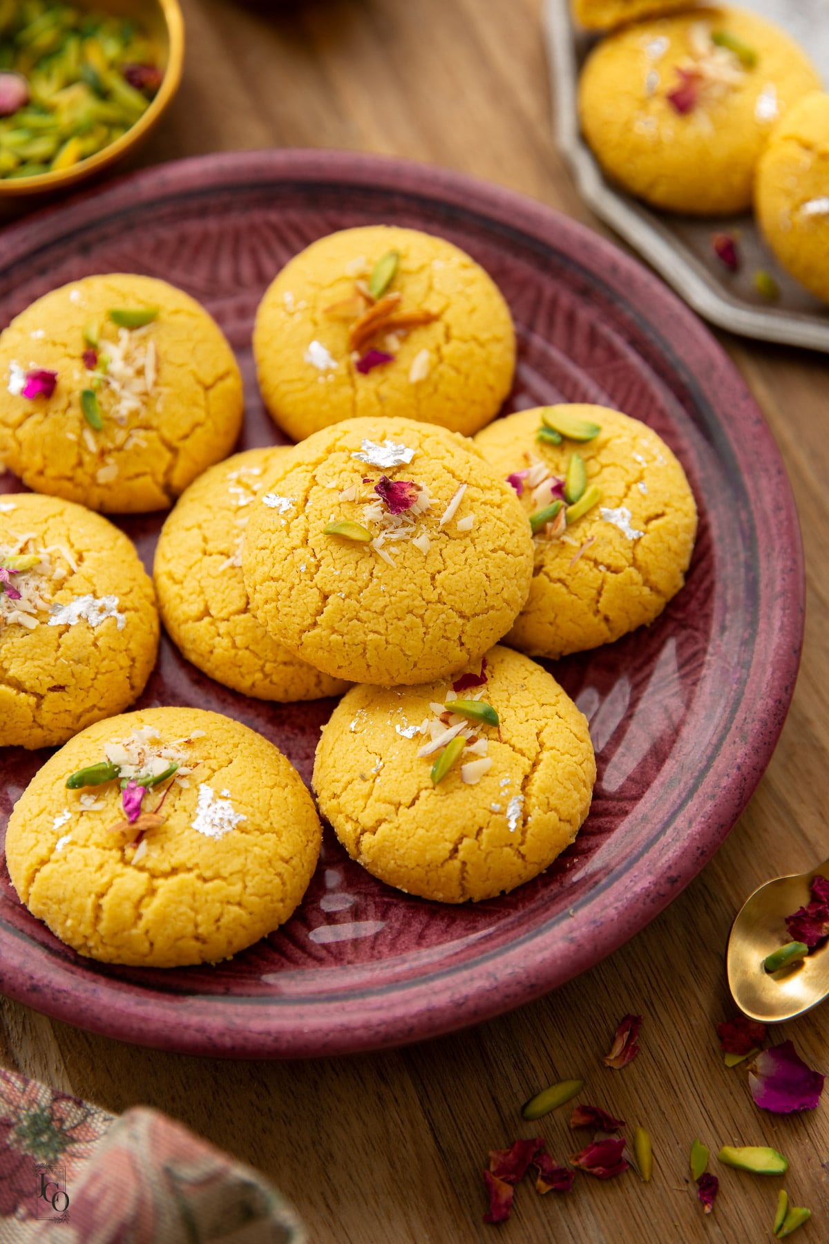 A cluster of crackly, melt-in-the-mouth besan nankhatai cookies topped with slivered nuts and edible flower petals, served on a textured maroon plate.