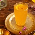 Orange colour saffron cardamom lemonade in a glass, topped with lemon slices and a few strands of saffron.