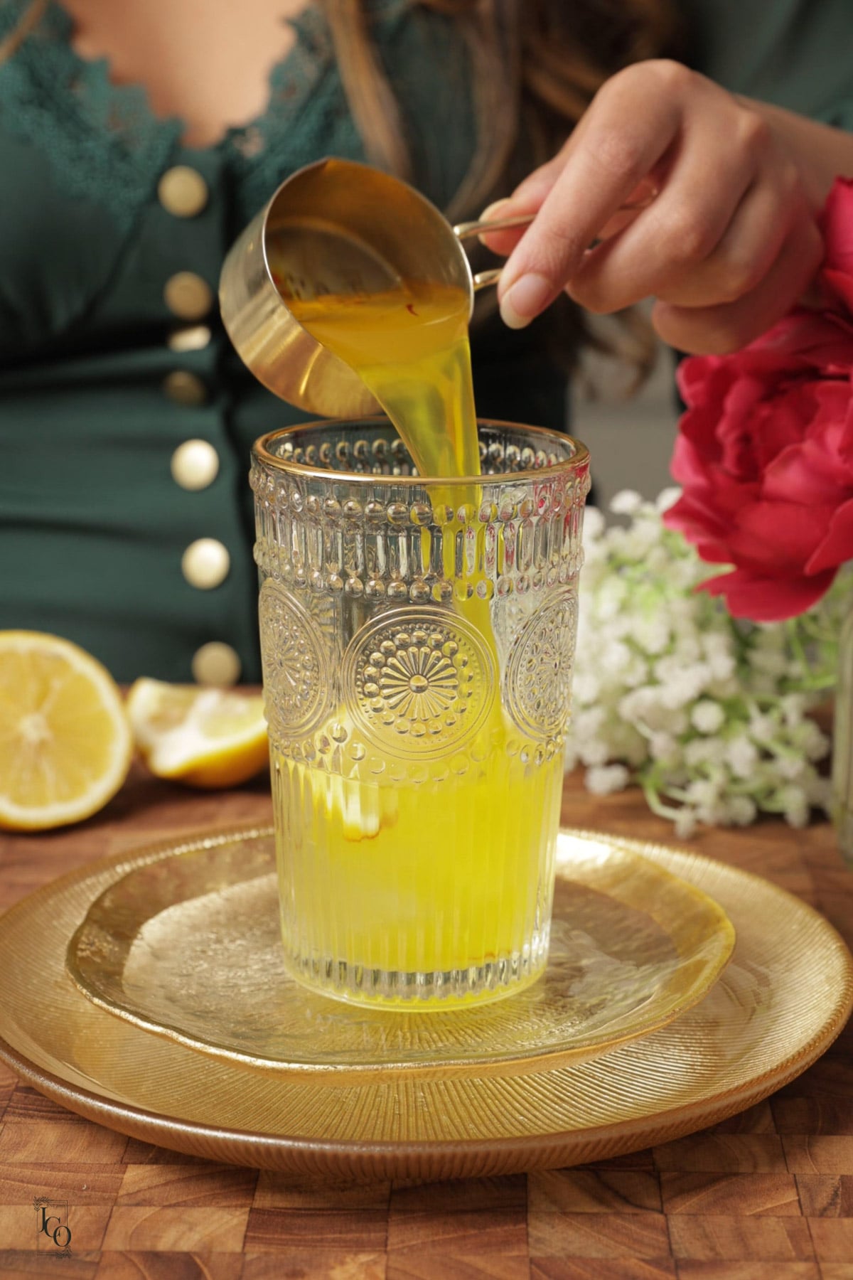 A female hand is pouring saffron cardamom syrup from cup into glass over the ice cubes.