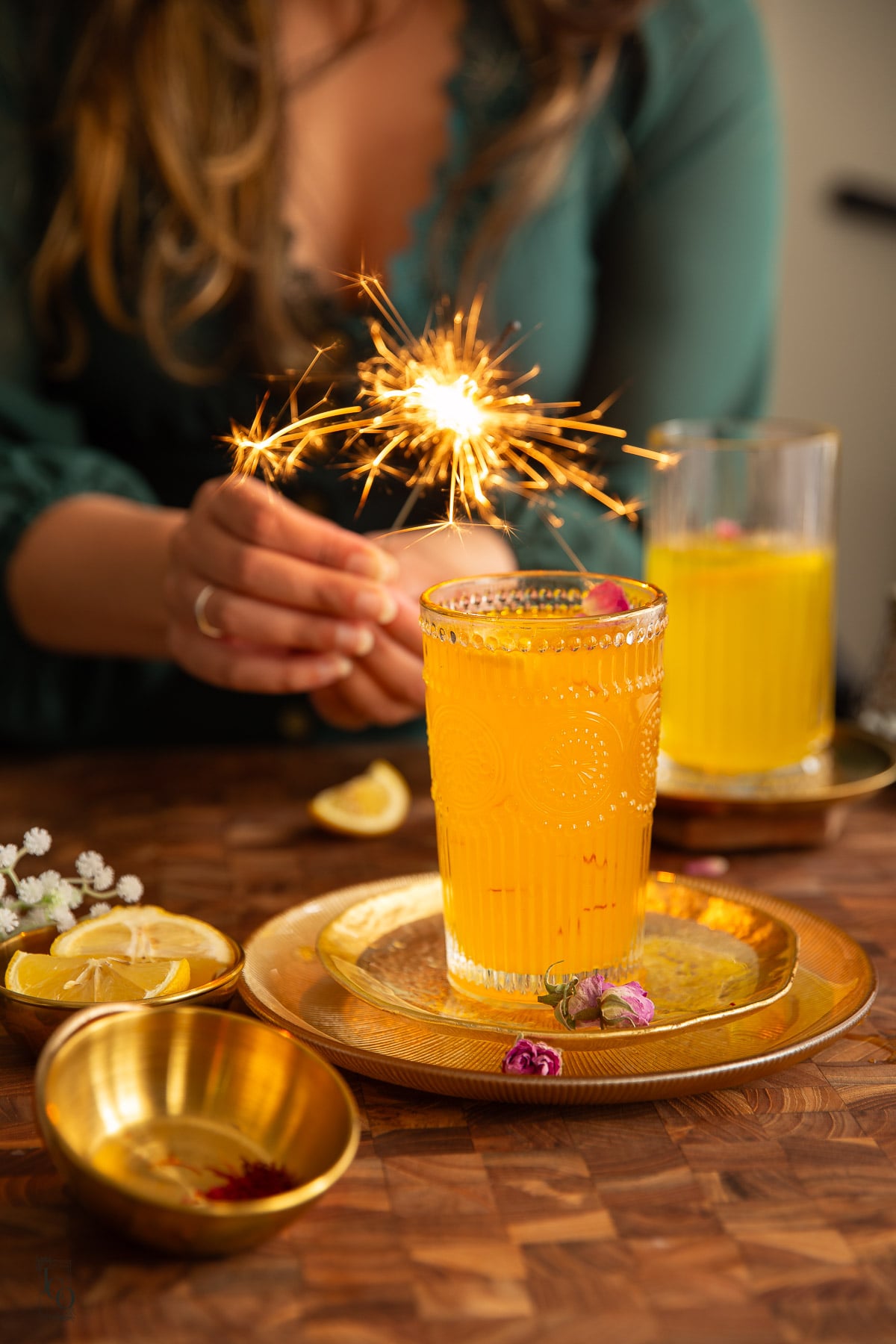 Two saffron cardamom glasses placed on gold serving tray, in the background a female hand is holding a lit sparkler.